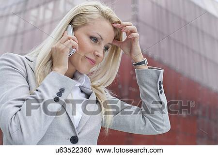 Stock Image - Close-up of confused businesswoman communicating on cell phone against office building. Fotosearch