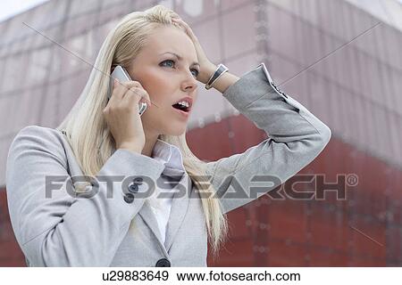 Stock Photo - Close-up of shocked businesswoman communicating on cell phone against office building. Fotosearch