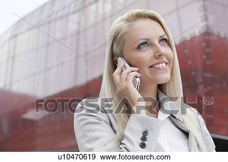 Close-up of smiling businesswoman conversing on cell phone against office building View Large Photo Image Stock Photo - Close-up of smiling businesswoman conversing on cell phone against office building. Fotosearch