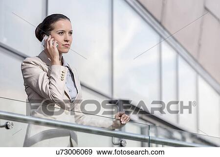 Confident young businesswoman using smart phone at office railing View Large Photo Image Stock Photo - Confident young businesswoman using smart phone at office railing. Fotosearch