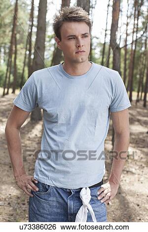 Stock Photograph - Confident young man with hands on hips in forest. Fotosearch