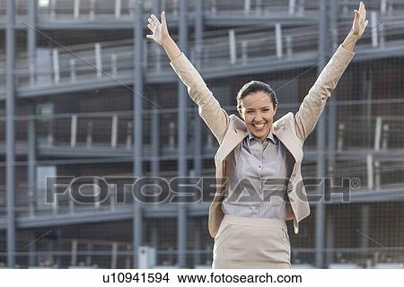 Picture - Excited young businesswoman with arms raised standing against office building. Fotosearch