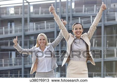 Stock Image - Excited young businesswomen gesturing thumbs up against office building. Fotosearch
