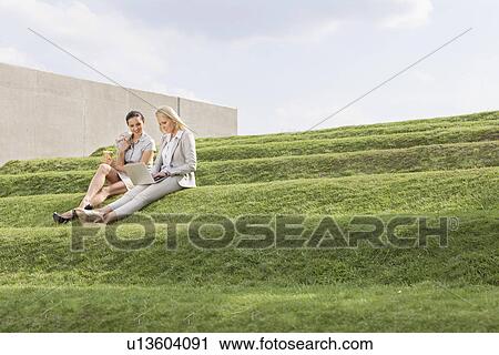 Stock Image - Full length of beautiful young businesswomen using laptop while sitting on grass steps against sky. Fotosearch
