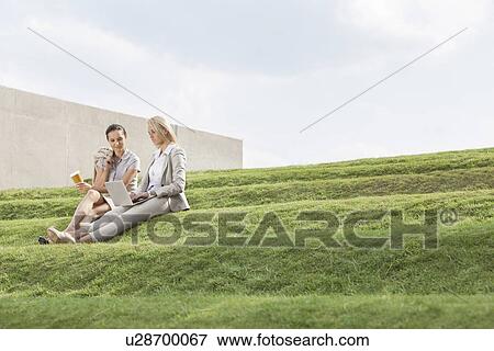 Full length of businesswomen with disposable coffee cup and laptop sitting on grass steps against sky View Large Photo Image Stock Photo - Full length of businesswomen with disposable coffee cup and laptop sitting on grass steps against sky. Fotosearch