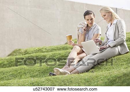 Full length of businesswomen with disposable coffee cup looking at laptop while sitting on grass steps View Large Photo Image Stock Photo - Full length of businesswomen with disposable coffee cup looking at laptop while sitting on grass steps. Fotosearch