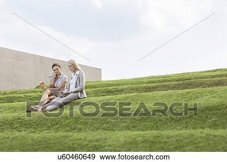 Full length of female business executives with disposable coffee cup and laptop sitting on grass steps against sky View Large Photo Image Stock Photo - Full length of female business executives with disposable coffee cup and laptop sitting on grass steps against sky. Fotosearch