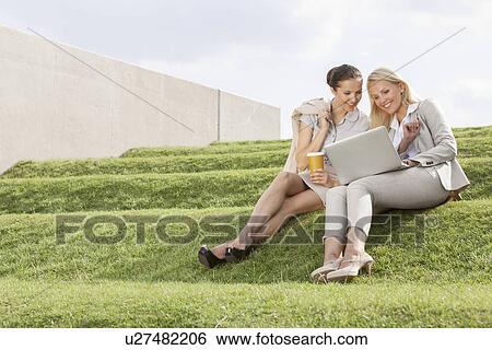 Stock Photograph - Full length of happy businesswomen looking at laptop while sitting on grass steps. Fotosearch