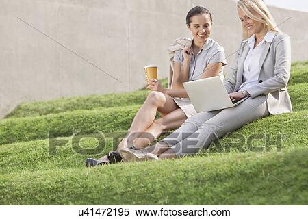 Stock Photography - Full length of happy businesswomen looking at laptop while sitting on grass steps against wall. Fotosearch