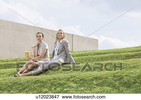 Stock Photo - Full length of relaxed businesswomen in formals sitting on grass steps against sky. Fotosearch