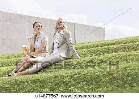 Stock Photo - Full length of relaxed businesswomen sitting on grass steps against sky. Fotosearch