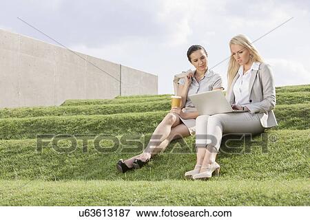 Full length of serious businesswomen looking at laptop while sitting on grass steps against sky View Large Photo Image Stock Photo - Full length of serious businesswomen looking at laptop while sitting on grass steps against sky. Fotosearch
