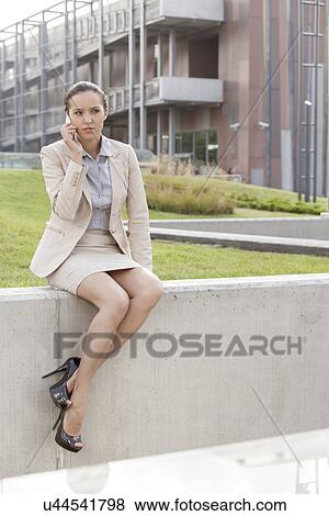 Full length of young businesswoman using mobile phone while sitting on wall against office building View Large Photo Image Stock Photo - Full length of young businesswoman using mobile phone while sitting on wall against office building. Fotosearch