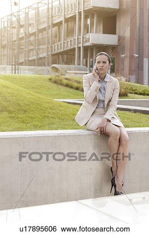 Stock Photograph - Full length of young businesswoman using cell phone while sitting on wall against office building. Fotosearch