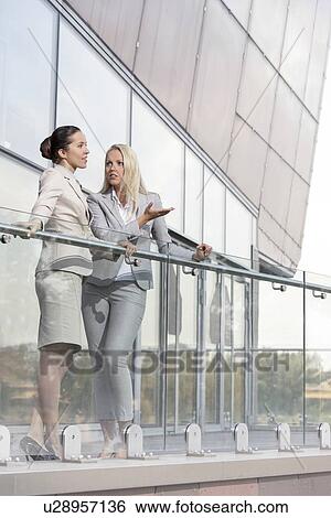 Stock Photograph - Full length of young businesswomen conversing at office balcony. Fotosearch