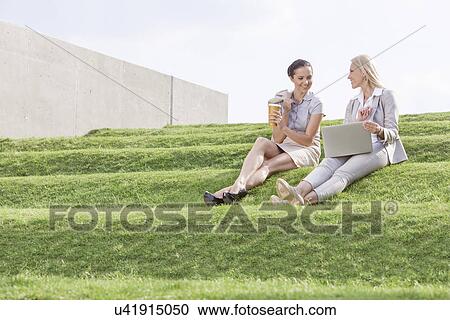 Full length of young businesswomen with disposable coffee cup and laptop sitting on grass steps against sky View Large Photo Image Stock Image - Full length of young businesswomen with disposable coffee cup and laptop sitting on grass steps against sky. Fotosearch