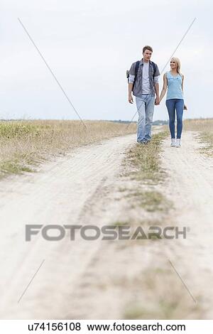 Stock Photo - Full length of young hiking couple walking on trail at field. Fotosearch