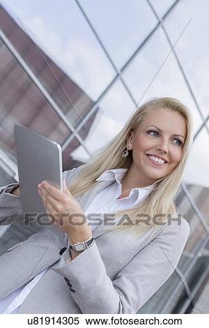Stock Photography - Happy businesswoman looking away while holding digital tablet against office building. Fotosearch