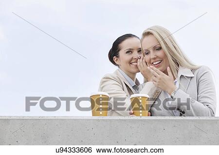 Happy businesswomen with disposable coffee cups sharing secrets against clear sky View Large Photo Image Stock Photograph - Happy businesswomen with disposable coffee cups sharing secrets against clear sky. Fotosearch