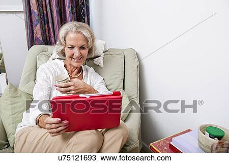 Stock Image - Happy senior woman looking at digital tablet on armchair in house. Fotosearch