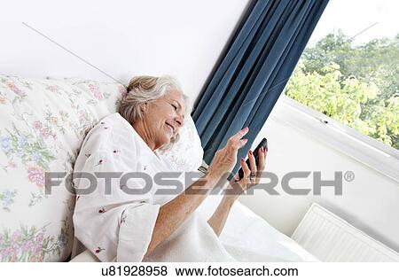 Happy senior woman using cell phone in bed at home View Large Photo Image Stock Photo - Happy senior woman using cell phone in bed at home. Fotosearch