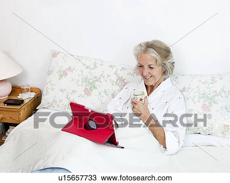 Happy senior woman using digital tablet while having coffee on bed at home View Large Photo Image Stock Image - Happy senior woman using digital tablet while having coffee on bed at home. Fotosearch