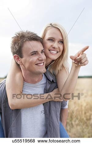 Happy woman showing something to man while enjoying piggyback ride at field View Large Photo Image Stock Photo - Happy woman showing something to man while enjoying piggyback ride at field. Fotosearch