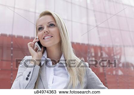 Stock Image - Happy young businesswoman communicating on smart phone against office building. Fotosearch