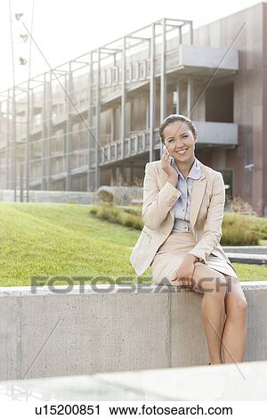 Happy young businesswoman using mobile phone while sitting on wall against office building View Large Photo Image Stock Image - Happy young businesswoman using mobile phone while sitting on wall against office building. Fotosearch