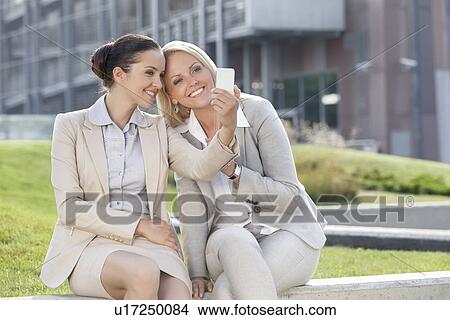 Happy young businesswomen taking self portrait through mobile phone against office building View Large Photo Image Picture - Happy young businesswomen taking self portrait through mobile phone against office building. Fotosearch