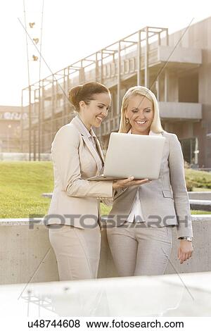 Stock Photo - Happy young businesswomen using laptop together while standing against building. Fotosearch