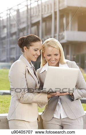 Happy young businesswomen using laptop together against building View Large Photo Image Stock Photo - Happy young businesswomen using laptop together against building. Fotosearch