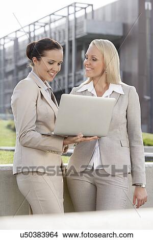 Happy young businesswomen with laptop standing against office building View Large Photo Image Picture - Happy young businesswomen with laptop standing against office building. Fotosearch