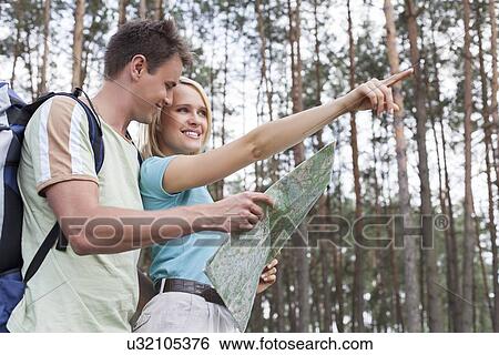 Stock Photograph - Happy young hiking couple holding map with woman pointing away in forest. Fotosearch