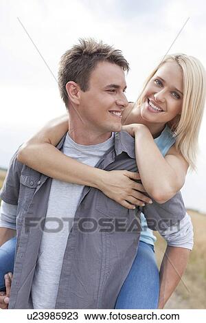 Stock Image - Happy young man giving piggyback ride to woman on field. Fotosearch