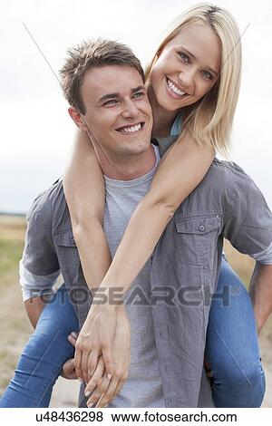 Stock Photo - Happy young man giving piggyback ride to girlfriend on field. Fotosearch