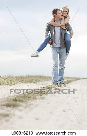 Stock Photo - Happy young man giving piggyback ride to woman on trail at field. Fotosearch