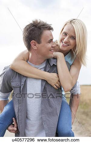 Happy young man giving piggyback ride to woman on field View Large Photo Image Stock Photograph - Happy young man giving piggyback ride to woman on field. Fotosearch