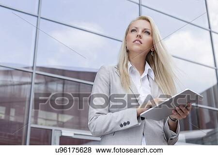 Low angle view of businesswoman holding digital tablet while looking away against office building View Large Photo Image Stock Photo - Low angle view of businesswoman holding digital tablet while looking away against office building. Fotosearch