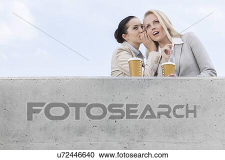 Low angle view of businesswoman whispering in coworker's ear while standing on terrace against sky View Large Photo Image Stock Image - Low angle view of businesswoman whispering in coworker's ear while standing on terrace against sky. Fotosearch