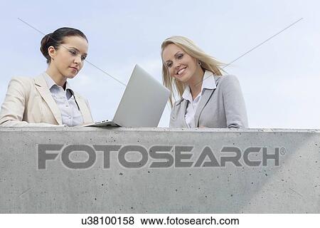 Low angle view of businesswoman using laptop while standing with colleague on terrace against sky View Large Photo Image Stock Photo - Low angle view of businesswoman using laptop while standing with colleague on terrace against sky. Fotosearch