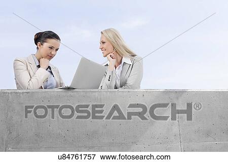 Stock Photo - Low angle view of concentrated businesswoman using laptop while standing with happy coworker on terrace against sky. Fotosearch