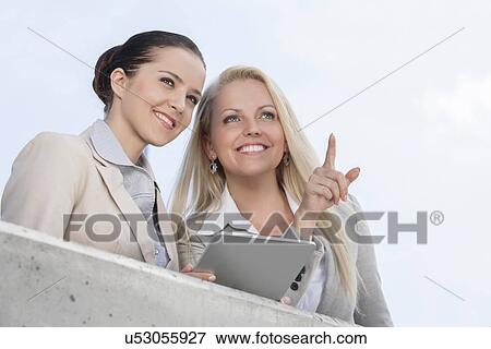 Stock Photo - Low angle view of happy businesswoman pointing while standing with coworker on terrace against sky. Fotosearch