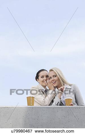 Stock Photo - Low angle view of happy businesswoman whispering in coworker's ear while standing on terrace against sky. Fotosearch