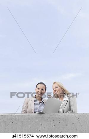 Picture - Low angle view of happy young businesswomen with laptop looking away while standing on terrace against sky. Fotosearch