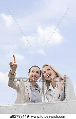 Stock Photo - Low angle view of happy young businesswoman showing something to coworker while standing on terrace against sky. Fotosearch