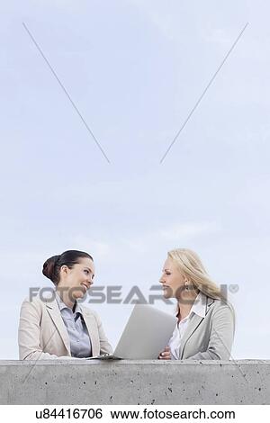 Low angle view of happy young businesswomen with laptop discussing while standing on terrace against sky View Large Photo Image Stock Photograph - Low angle view of happy young businesswomen with laptop discussing while standing on terrace against sky. Fotosearch