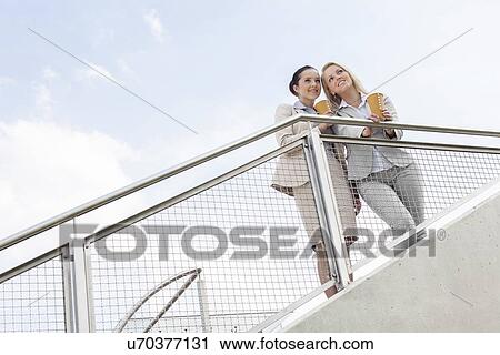 Low angle view of young businesswomen with disposable coffee cups standing by railing against sky View Large Photo Image Stock Image - Low angle view of young businesswomen with disposable coffee cups standing by railing against sky. Fotosearch
