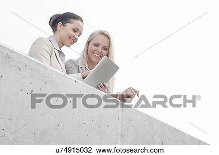 Stock Image - Low angle view of young businesswomen using digital tablet on office terrace against sky. Fotosearch