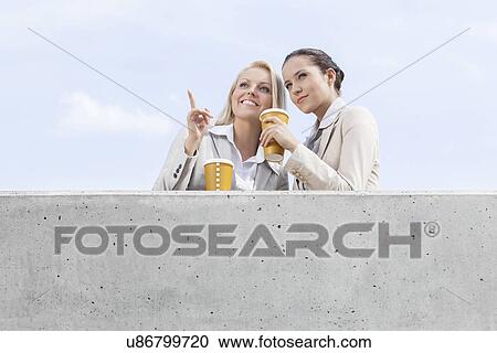 Stock Image - Low angle view of young businesswomen with disposable coffee cups looking away while standing on terrace against sky. Fotosearch